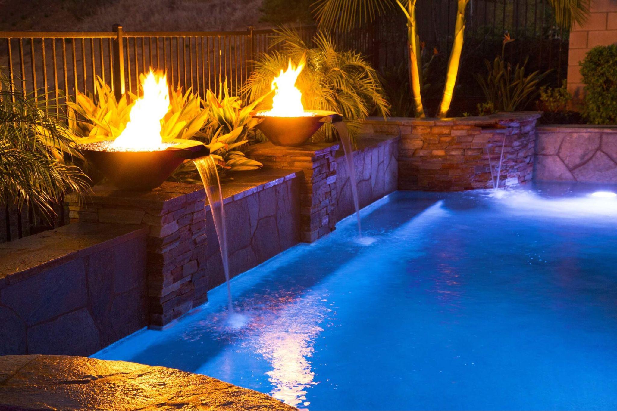 Modern pool with glowing fire bowls and cascading water features illuminated by soft evening lighting.