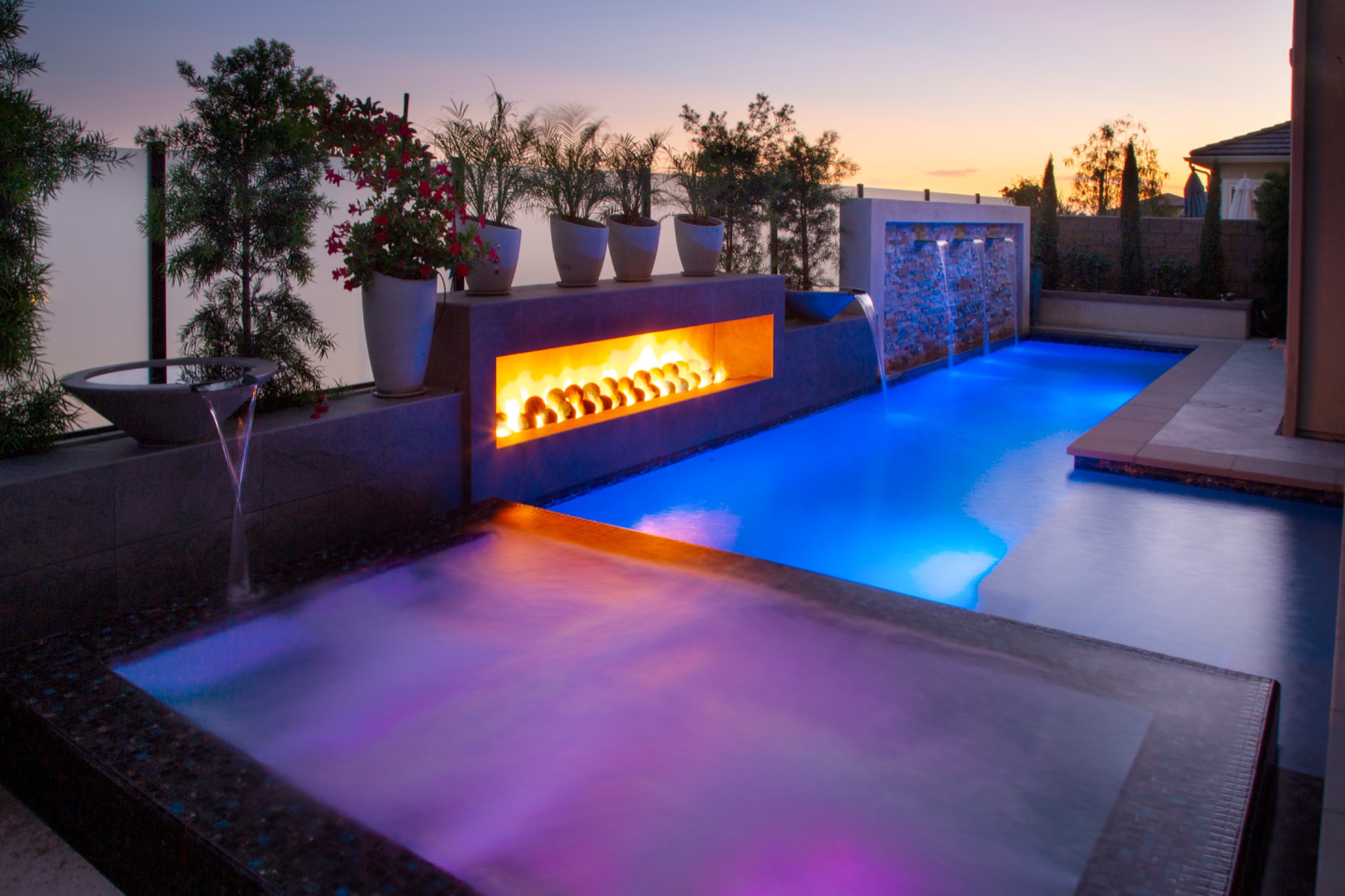 Luxury inground swimming pool with color-changing LED lights, built-in linear fireplace, sheer descent water features, and an attached spa at twilight