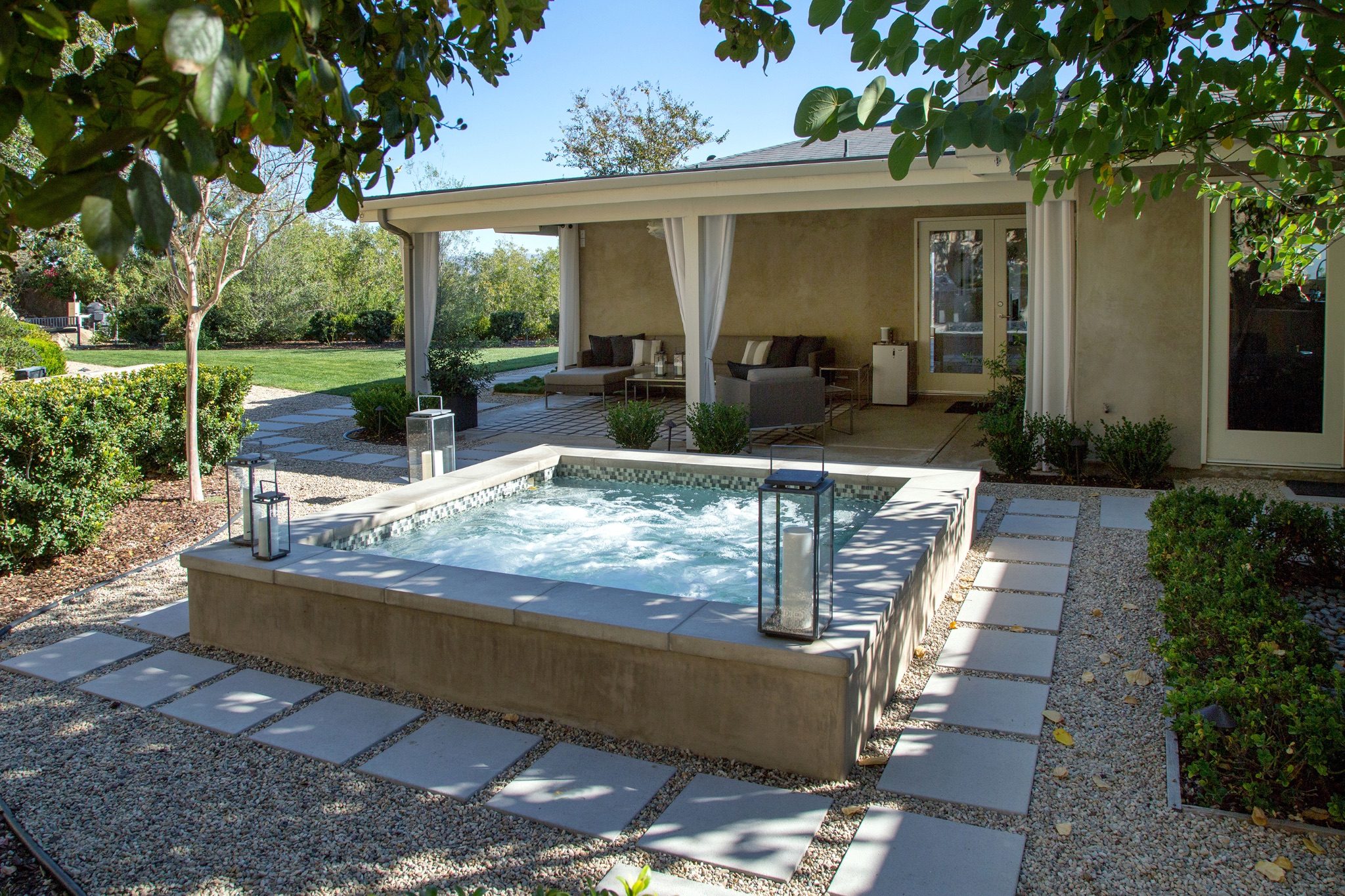 Raised concrete residential spa with bubbling water and mosaic tile trim set in a landscaped backyard with paver pathways, lanterns, and a covered patio lounge area