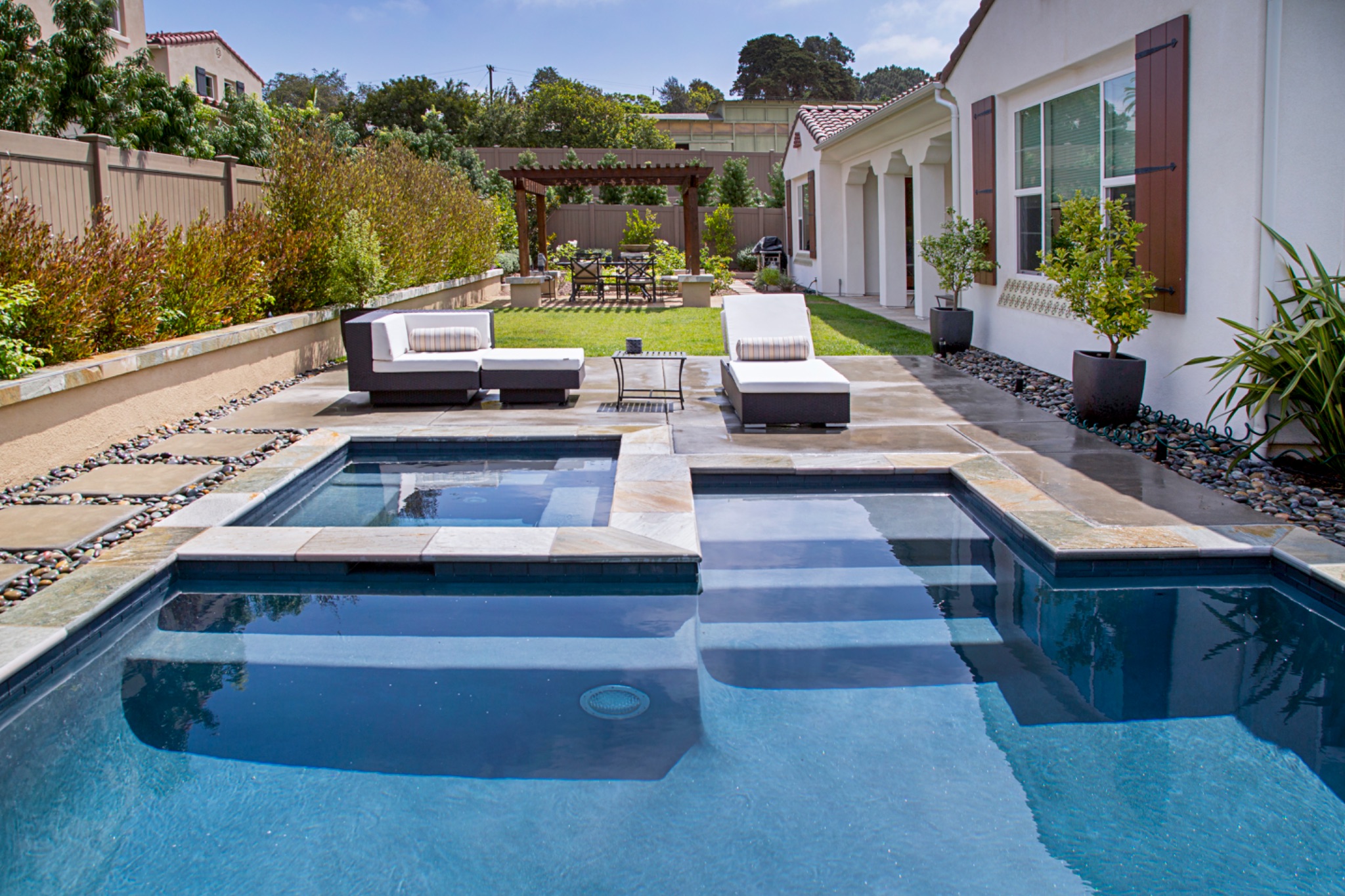 Rectangular inground swimming pool with an attached square spa, natural stone decking, outdoor lounge furniture, and a pergola in a Southern California backyard