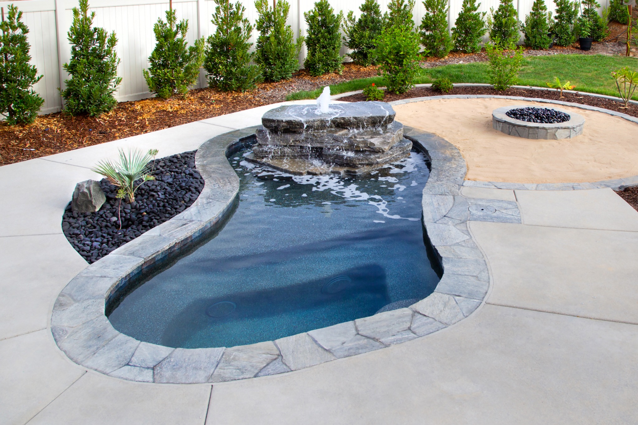 Clean freeform residential swimming pool with a tiered stone fountain, dark pebble accents, and an adjacent fire pit surrounded by landscaped hedges