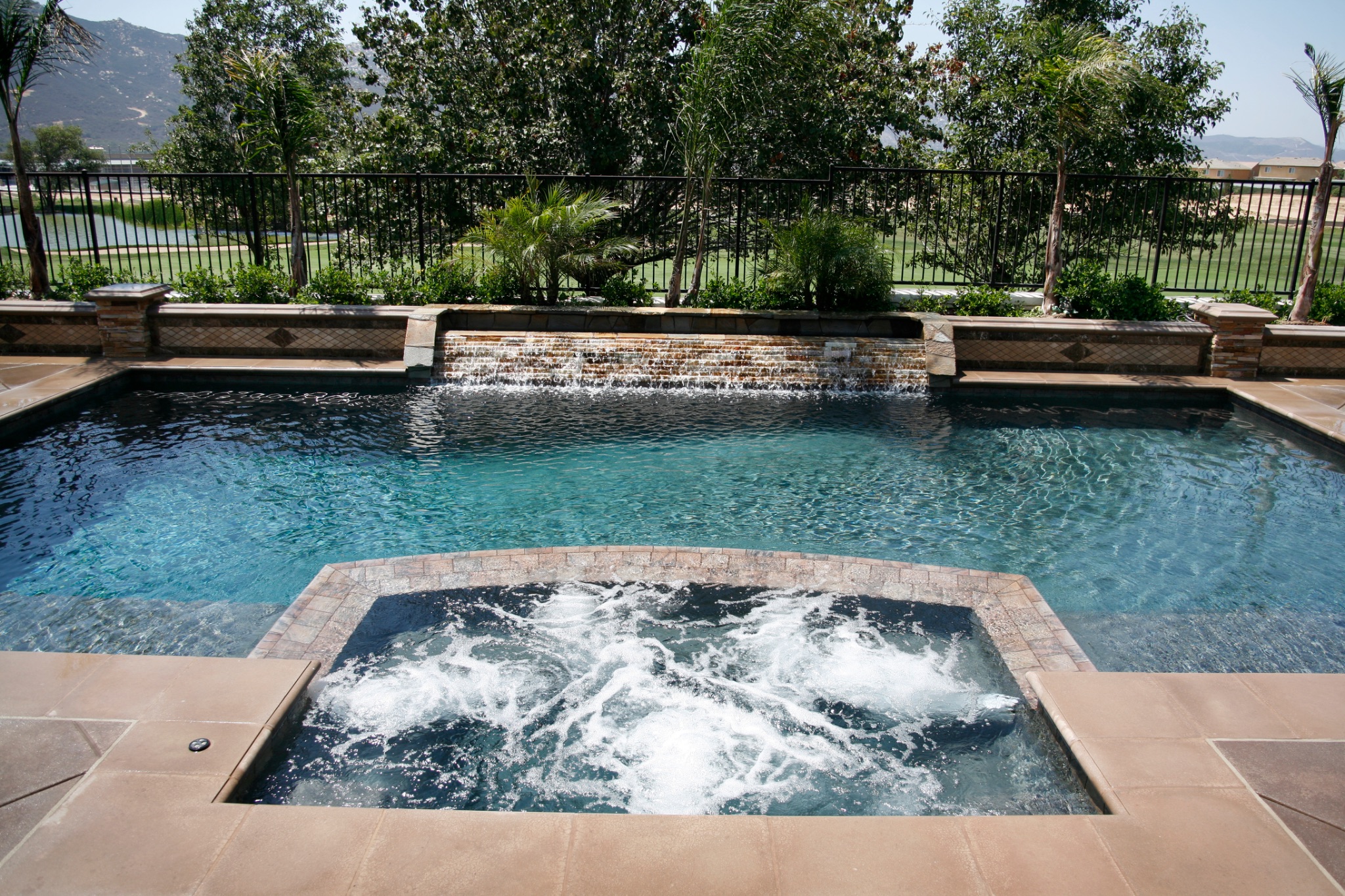 Inground swimming pool with a spillover spa, stacked stone waterfall feature, brick coping, and safety fencing overlooking a golf course in Southern California