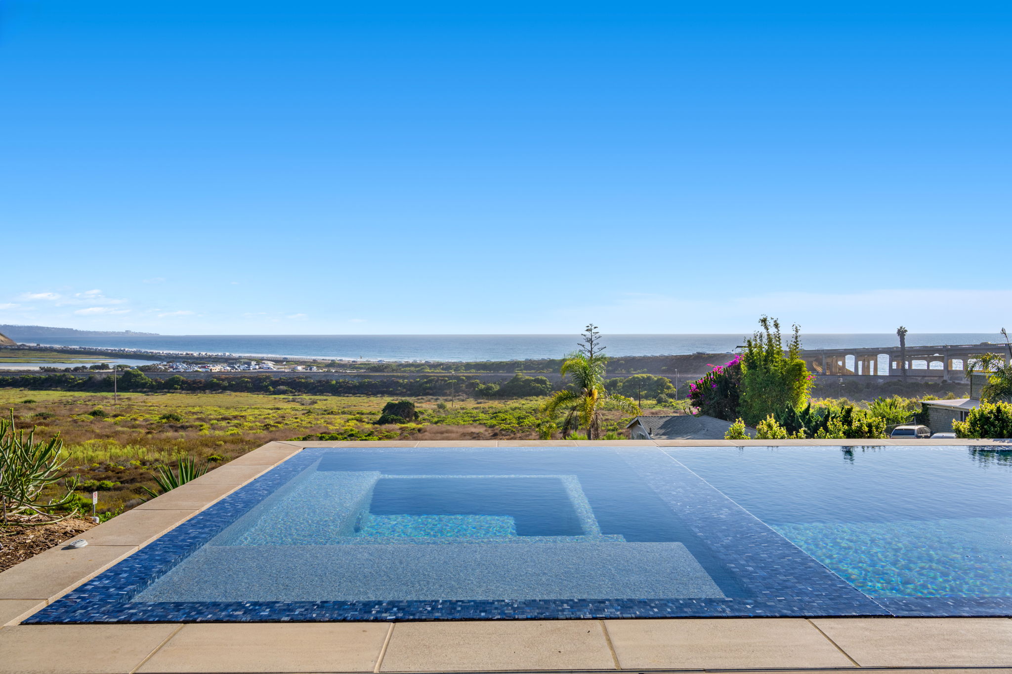Residential swimming pool with an integrated raised spa featuring blue mosaic tile, set on a hilltop patio with panoramic ocean views