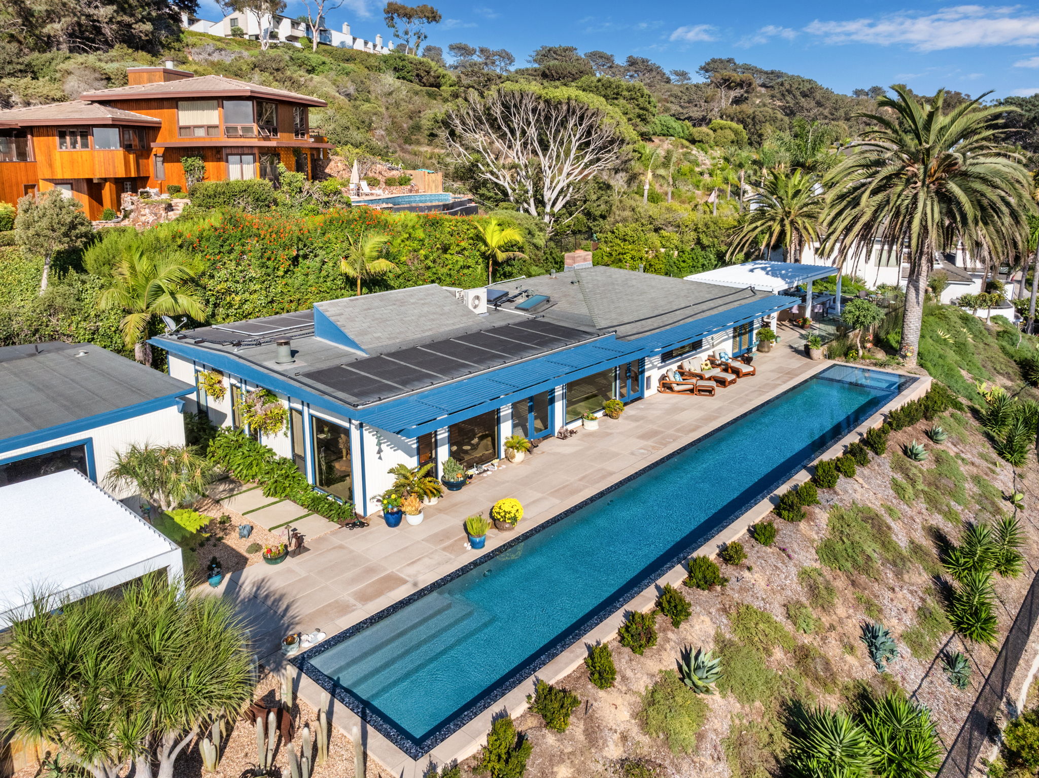 Aerial view of a long residential lap pool with dark tile finish running alongside a hillside home with solar panels, lounge chairs, and tropical landscaping